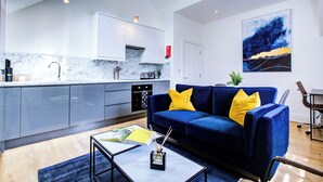 A modern kitchen with grey cabinets, a marble backsplash, and a blue sofa with yellow cushions. - Hybrid Resi - New Malden | 1 Bed Apartment (New Malden)