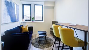 Een knus woongedeelte met een donkerblauwe bank, een klein zwart tafeltje en twee gele stoelen. Aan de muur hangt een groot abstract schilderij, en er is een raam met uitzicht op gebouwen.