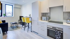 Een moderne keuken met witte kastjes, een magnetron en een oven. Er is een eethoek met een tafel en gele stoelen.