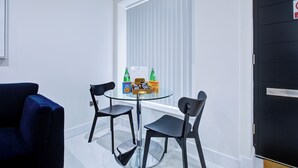 A room with a glass table, two black chairs, a blue sofa, and a window with blinds. - Hybrid Resi - Uxbridge|1 Bed Apartment (Uxbridge)