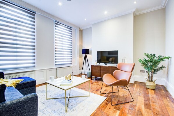 A modern living room with a sofa, a coffee table, a TV, and a potted plant. - Hybrid Resi - Battersea | 3 Bed Apartment (London)