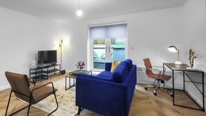 A modern living room with a blue sofa, a brown chair, a TV, and a desk with a lamp. - Hybrid Resi - Mitcham | 1 Bed Apartment (Mitcham)