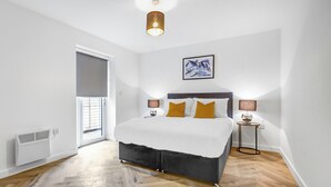 A bedroom with a bed, a bedside table, a lamp, a picture on the wall, and a window with blinds. - Hybrid Resi - Mitcham | 1 Bed Apartment (Mitcham)