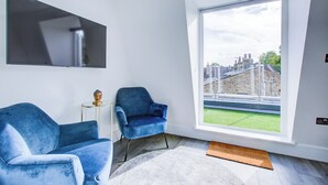 Ein modernes Wohnzimmer mit großem Fenster, einem Flachbildfernseher, zwei blauen Sessel und einem kleinen Beistelltisch.