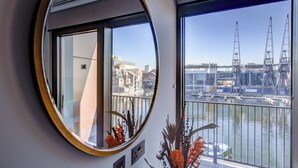 Quarto com uma grande janela que dá para uma marina com barcos e guindastes, um espelho redondo na parede e uma pequena planta no parapeito da janela.