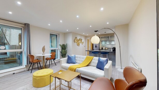 A modern living room with a sofa, ottoman, and coffee table, a dining area with chairs, and a kitchen in the background.