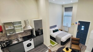 Une chambre d’hôtel compacte comprenant un coin cuisine, un lit avec une tête de lit grise, un petit coin salon et une porte avec une pancarte.