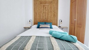 A neatly made bed with a striped blanket, two pillows, and a folded towel. There are two bedside tables with lamps, a wooden door, and a wardrobe.