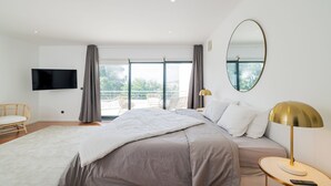 Una habitación de hotel moderna con una cama grande, un televisor de pantalla plana, un espejo redondo y un balcón con mobiliario exterior.