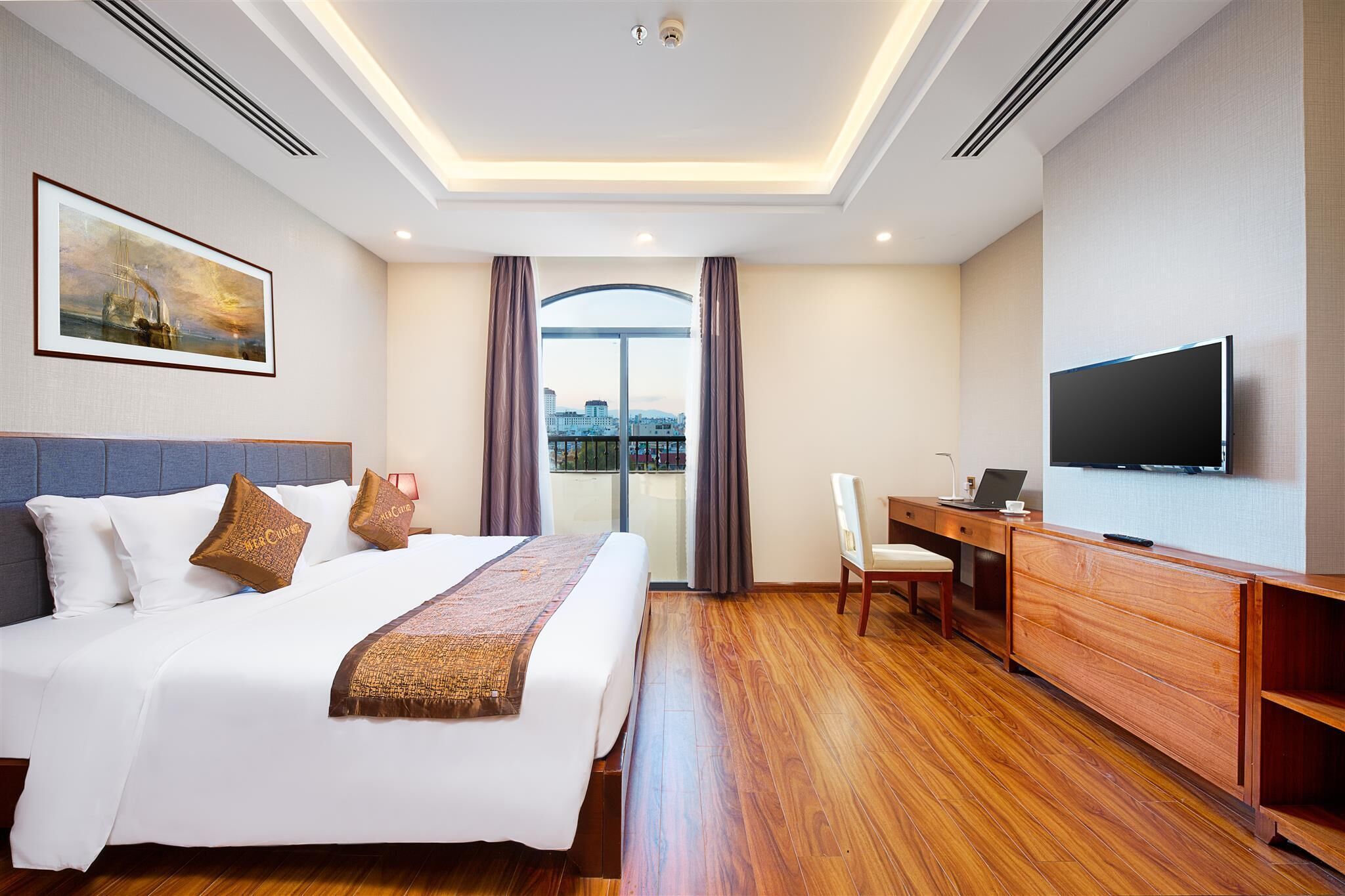 A hotel room with a bed, a desk, a television, and a view of the city through large windows.