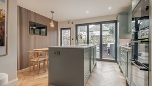 A modern kitchen with a central island, wooden dining table, and large sliding glass doors leading to a garden.