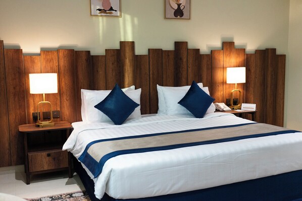 A neatly made bed with white and blue bedding, two bedside tables with lamps, and a wooden headboard. - Rayyan Al Janadriyah Serviced Apartments (Rumah)
