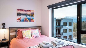 A bedroom with a large window, a bed with pink bedding, colorful pillows, a bedside lamp, and a painting of boats on a lake. - Revelstoke - Alpine Retreat (Revelstoke)