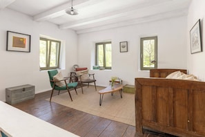 Una habitación con suelo de madera, una cama de madera, una silla verde, una mesa de madera y dos ventanas.