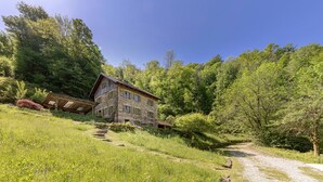 Une maison en pierre avec un toit en pente, entourée d’une végétation luxuriante et bordée d’un chemin de gravier.