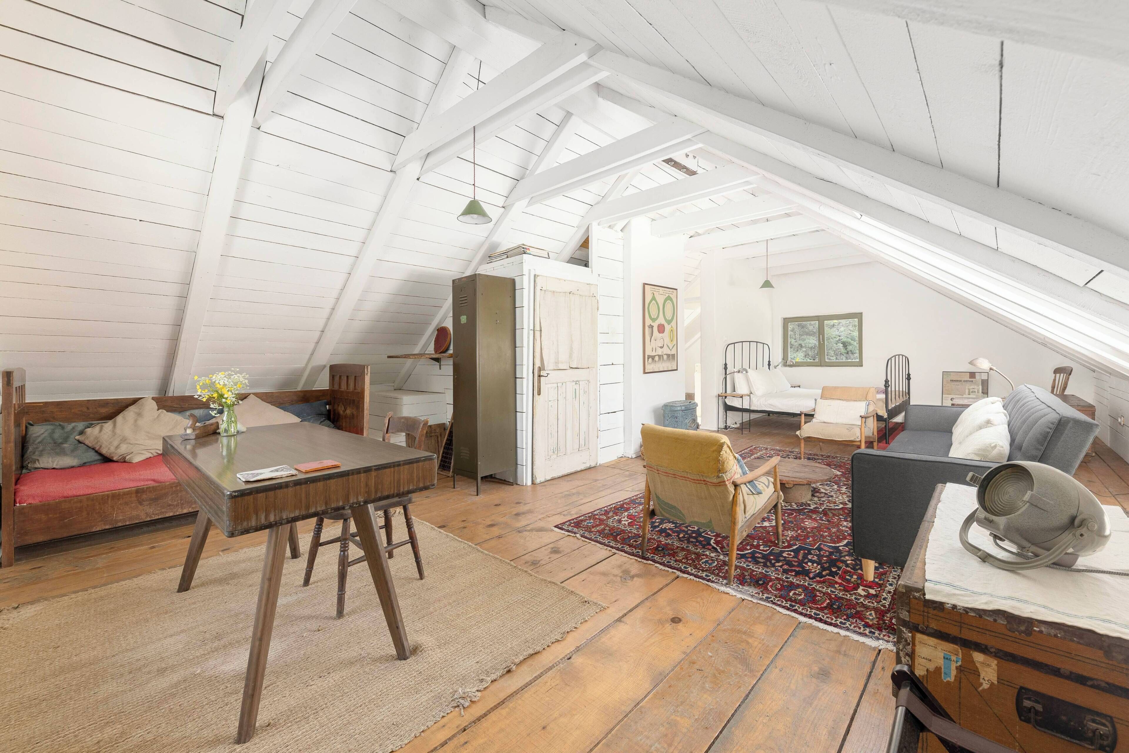 A loft-style room with a high ceiling, wooden beams, a dining table, a sofa, a chair, and a small table with a vase.