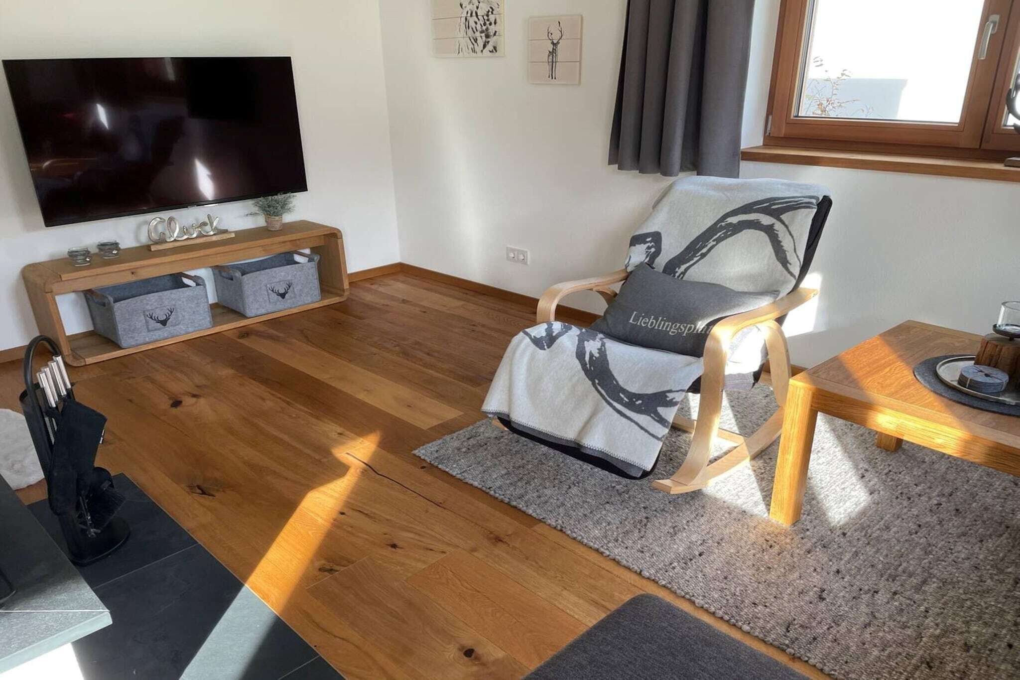 A modern living room with a flat-screen TV, a wooden coffee table, a grey armchair with a cushion, and a small plant on a wooden console table.