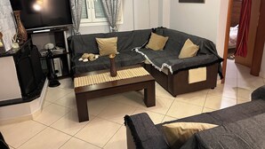 Een woonkamer met een donkergrijze bank, beige kussens, een bijzettafeltje, een televisie en een ingelijst schilderij aan de muur.