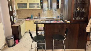 Een compacte keuken met gele kastjes, een roestvrijstalen koelkast en een glazen kast met keukengerei. Er staan twee zwarte barkrukken bij een klein eiland met een tijdschrift.