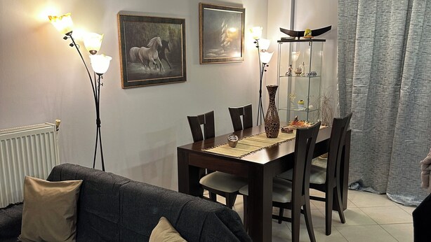 Een moderne woonkamer met een donkere bank, een eettafel met stoelen en een plafondventilator. Er zijn wandlampen en ingelijste foto's aan de muren.