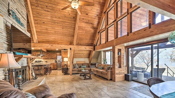 A spacious living area with a high ceiling, wooden beams, a fireplace, and a large window overlooking a scenic view. - Lakeside Haven with Sandy Beach, Sleeps 12, 3 Bath (Stigler)