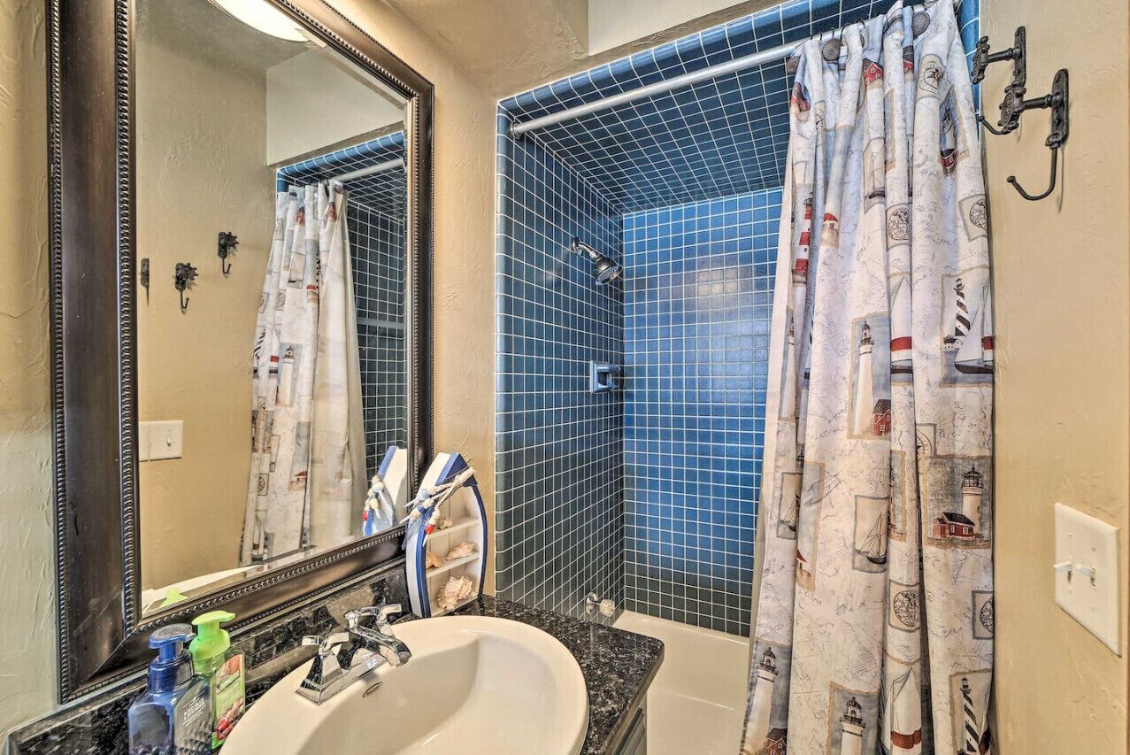 Un bany amb un mirall gran, un lavabo i una dutxera amb rajoles blaves i cortines amb temàtica marina.