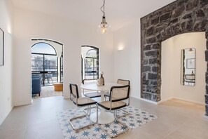 A dining area with a round table, four chairs, and a patterned rug. There is a stone wall and a window with a view of the outside.