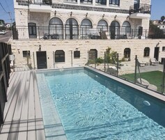Una piscina con muelle de madera, un edificio de piedra con ventanas arqueadas y mobiliario exterior.