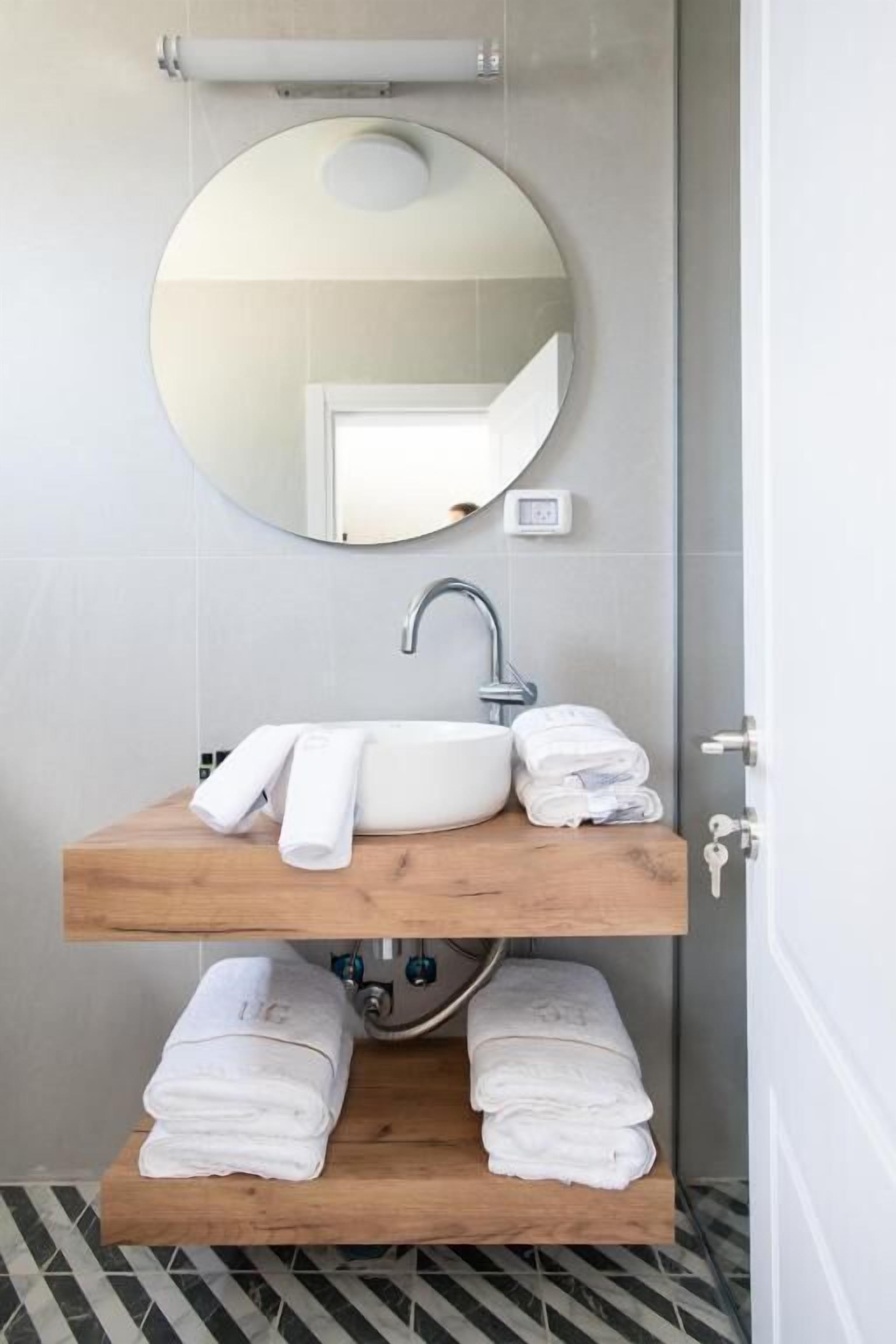 Une salle de bain avec un miroir rond, un lavabo blanc et deux étagères en bois où sont rangées des serviettes.