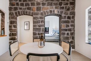 Comedor con una mesa redonda y cuatro sillas, una pared de piedra y vista hacia un dormitorio.