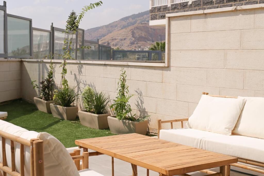 A rooftop patio with a wooden table and white sofa, surrounded by potted plants and a view of a mountain.