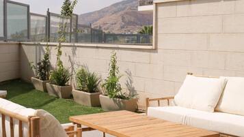 A rooftop patio with a wooden table and white sofa, surrounded by potted plants and a view of a mountain.
