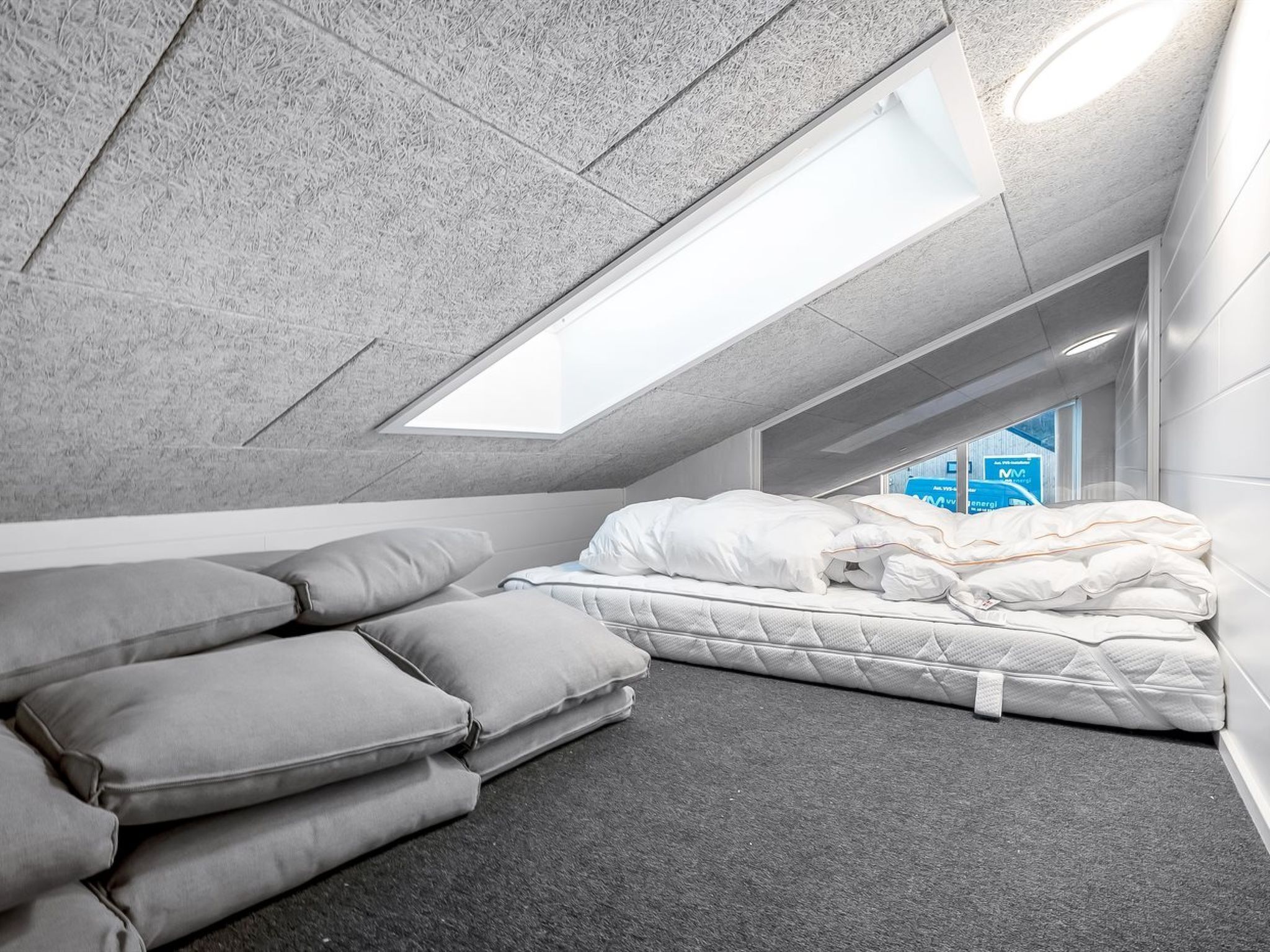 Habitació compacta i moderna amb sostre inclinat, un fanal i un llit ben fet amb roba de llit blanca, a més d'una pila de coixins grisos.