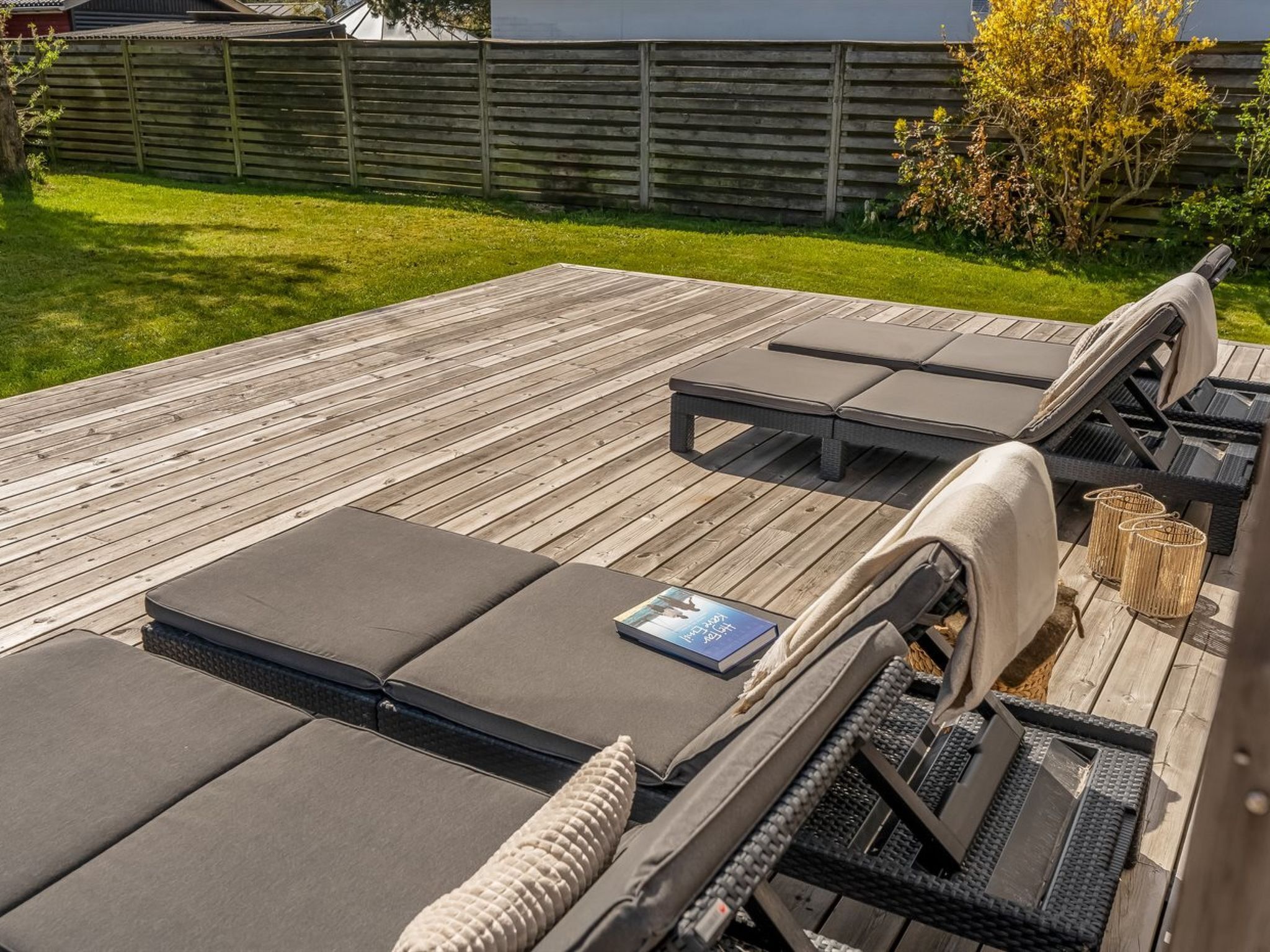A wooden deck with outdoor furniture including lounge chairs and a table, surrounded by a wooden fence and greenery.