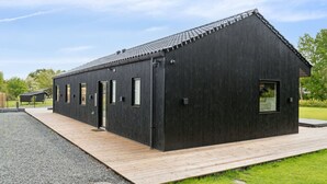 Un edificio moderno de color negro con techo inclinado, una terraza de madera y un camino de grava que conduce a él.