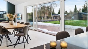 Un comedor moderno con una mesa grande y sillas, un jarrón con flores y vistas a un patio trasero con terraza de madera y vegetación.