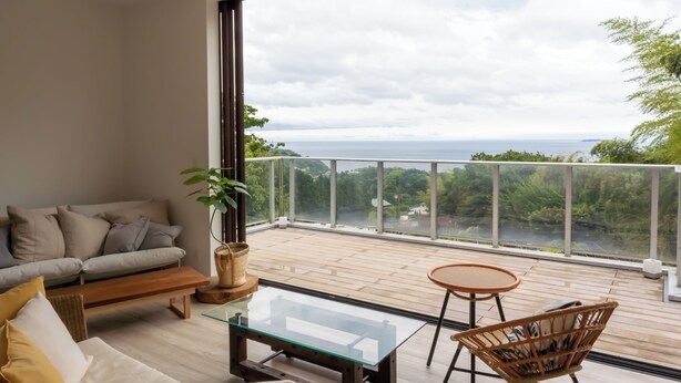 Un salón moderno con un sofá, una mesa de centro de cristal y una silla de mimbre. Un ventanal ofrece vistas a un balcón con suelo de madera y barandilla.