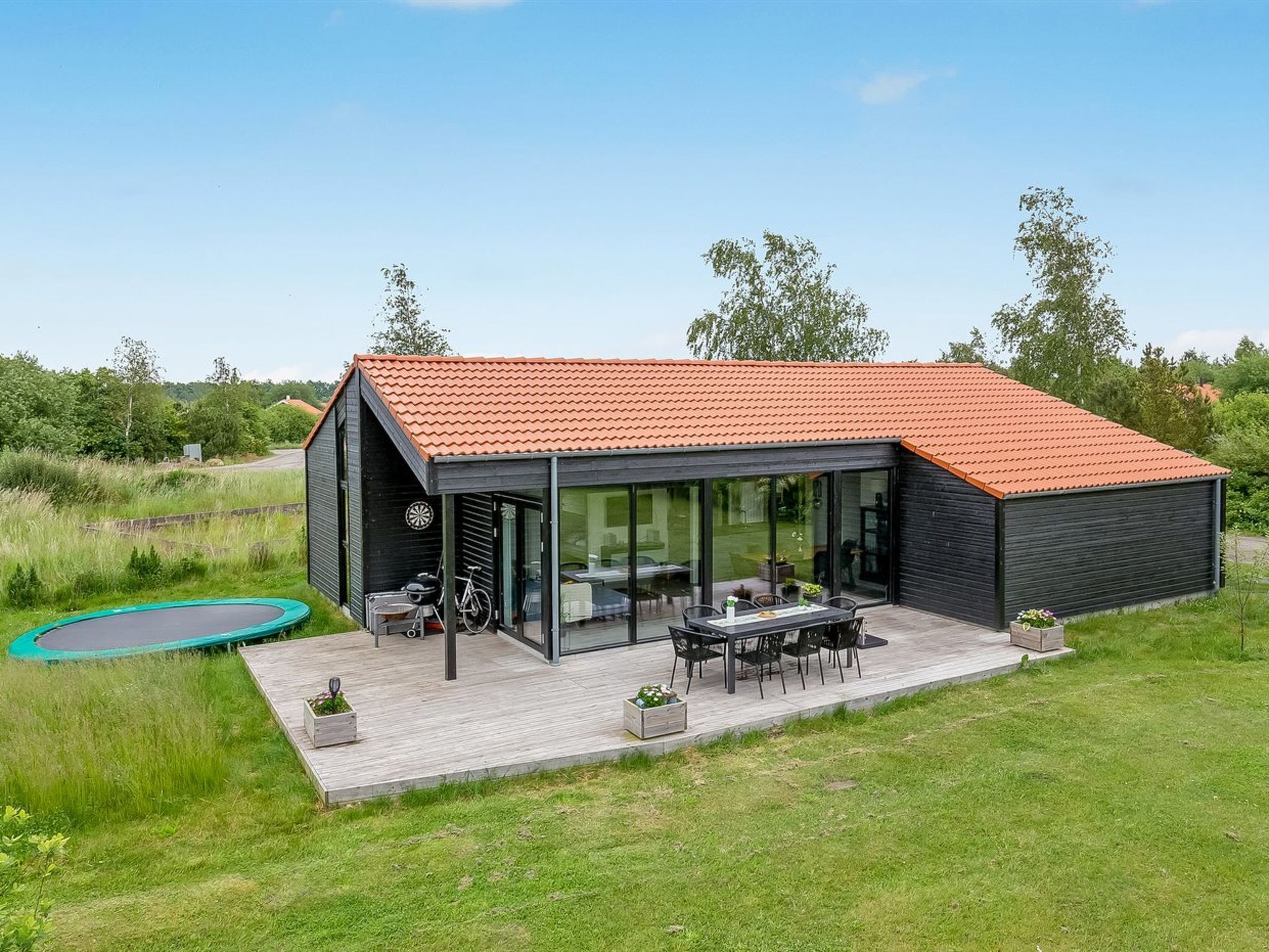 Une maison moderne avec un toit rouge, un patio fermé par des baies vitrées et un trampoline dans le jardin.