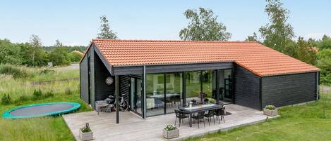 Une maison moderne avec un toit rouge, un patio fermé par des baies vitrées et un trampoline dans le jardin.
