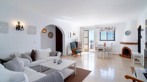 A spacious living room with a white sofa, a coffee table, and a fireplace. There is a dining area with a table and chairs.