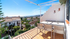 A balcony with a white table and wicker chairs, overlooking a cityscape with terracotta rooftops and greenery.
