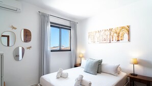 A hotel room with a bed, two bedside lamps, a window with curtains, a wall-mounted mirror, and a framed picture.