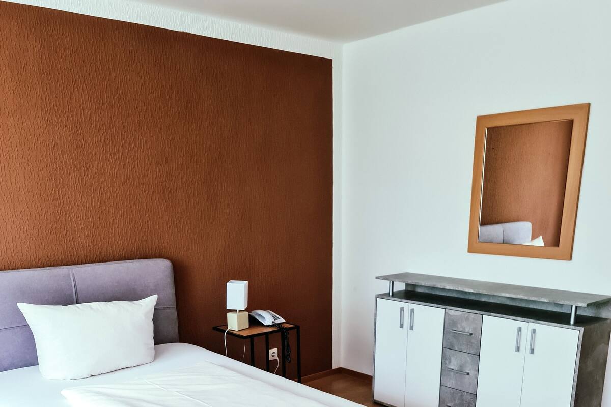 A modern hotel room with a large bed, a bedside table with a lamp, a wardrobe, and a mirror on the wall.