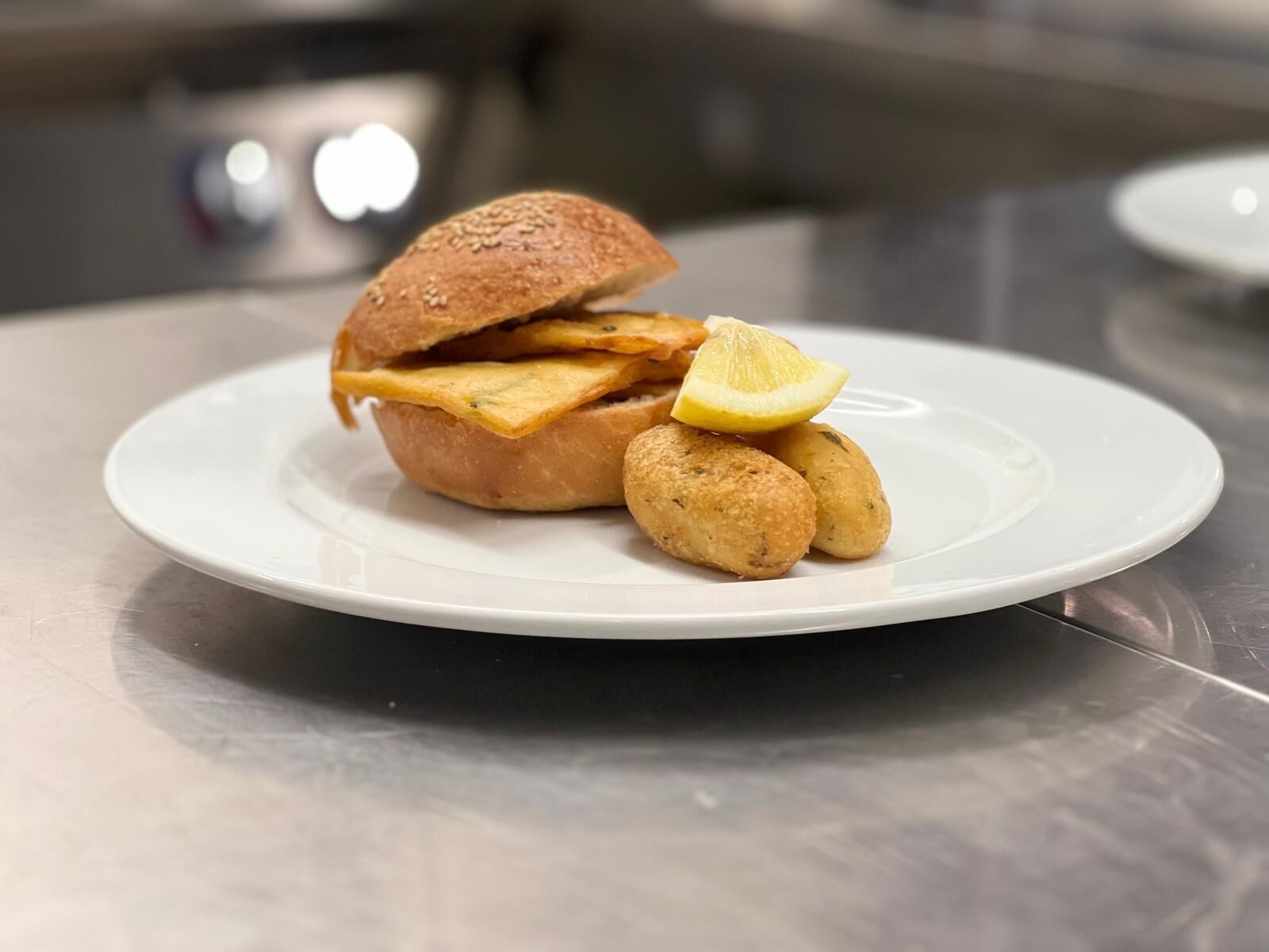 Um prato com sanduíche, acompanhamentos fritos e uma fatia de limão sobre uma mesa de aço inoxidável.