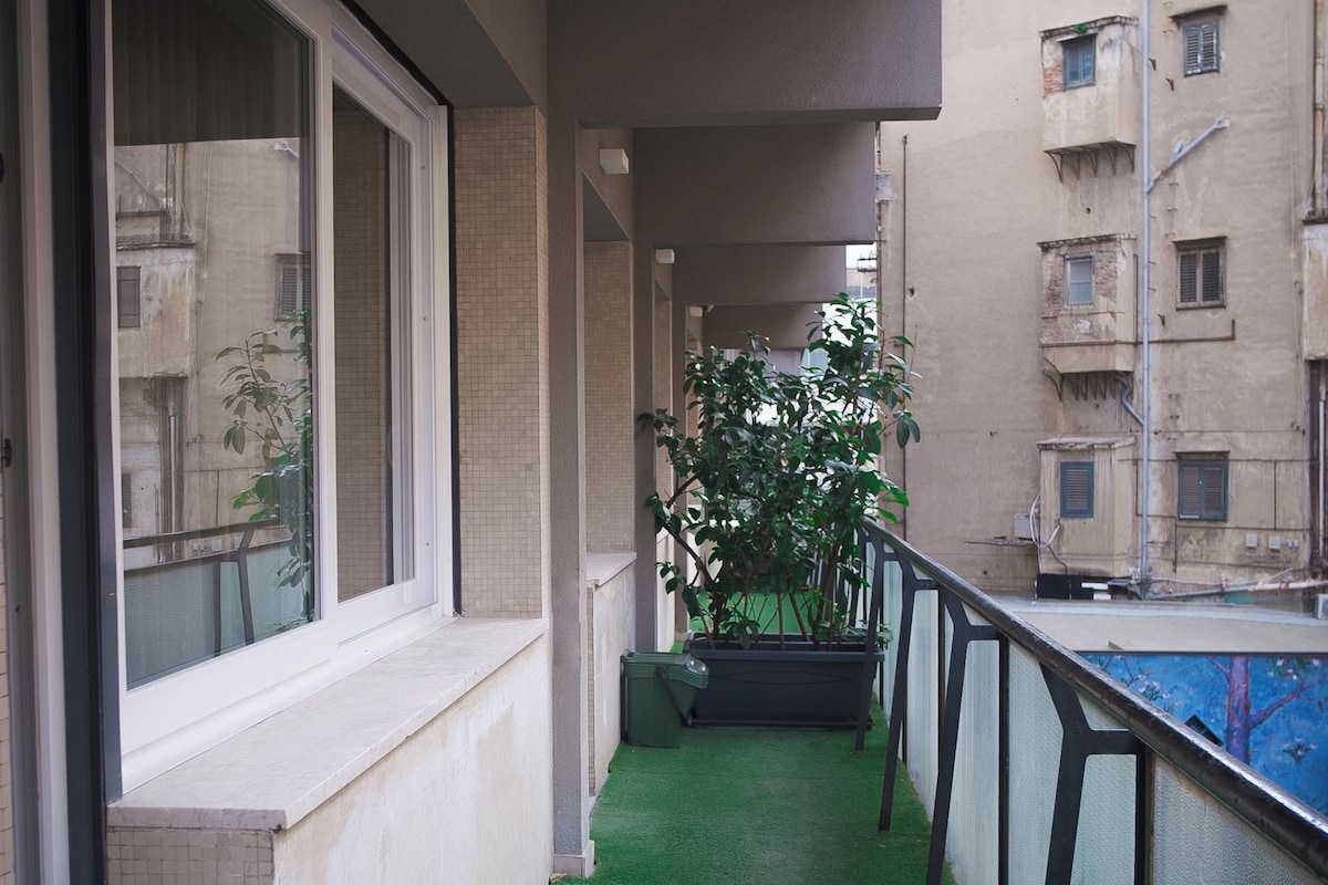 A narrow corridor with artificial green flooring, a window on the left, and a balcony on the right.