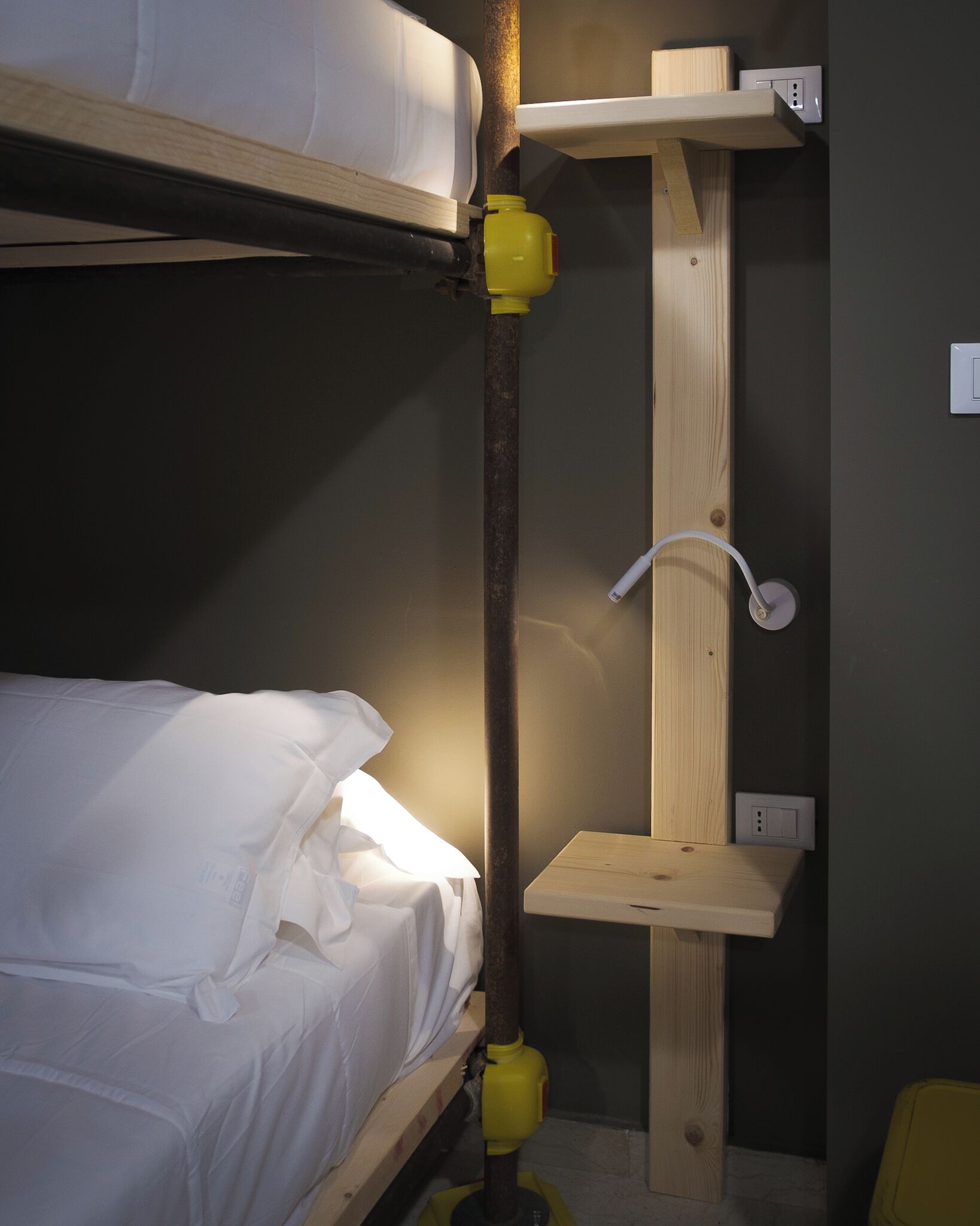 Un lit superposé avec une literie blanche, une étagère en bois et un objet jaune fixé au mur.