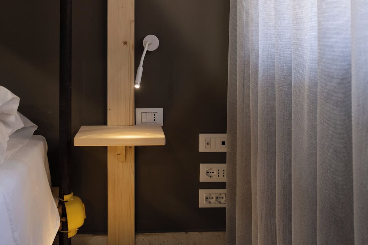 A compact bathroom with a wooden shelf, a white showerhead, and electrical outlets on the wall.