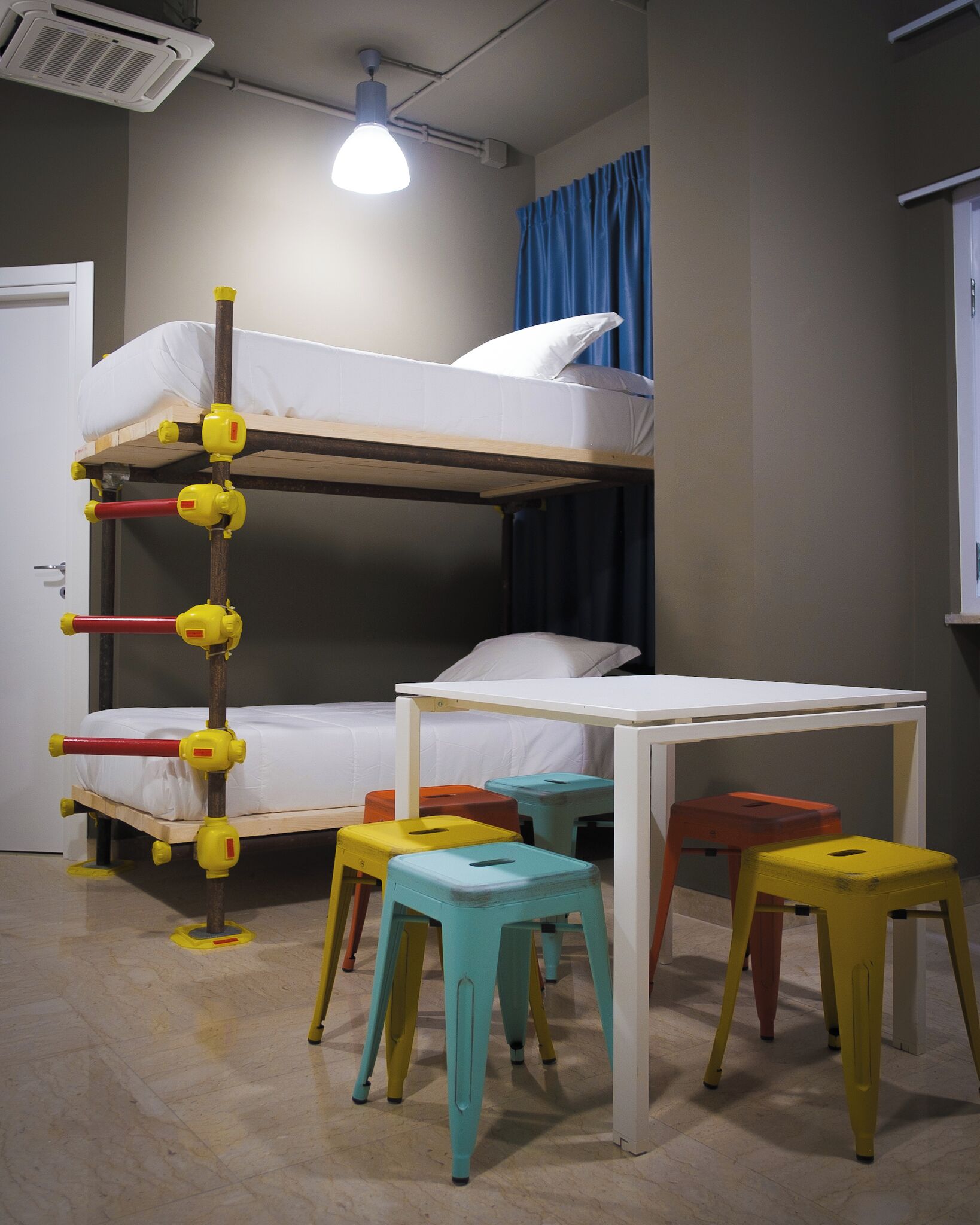 Uma cama beliche com escrivaninha e bancos num quarto com janela e cortinas.