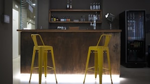A bar area with two yellow stools, a wooden counter, shelves with bottles, and a refrigerator. - Hostelò Luxury Hostel (Palermo)