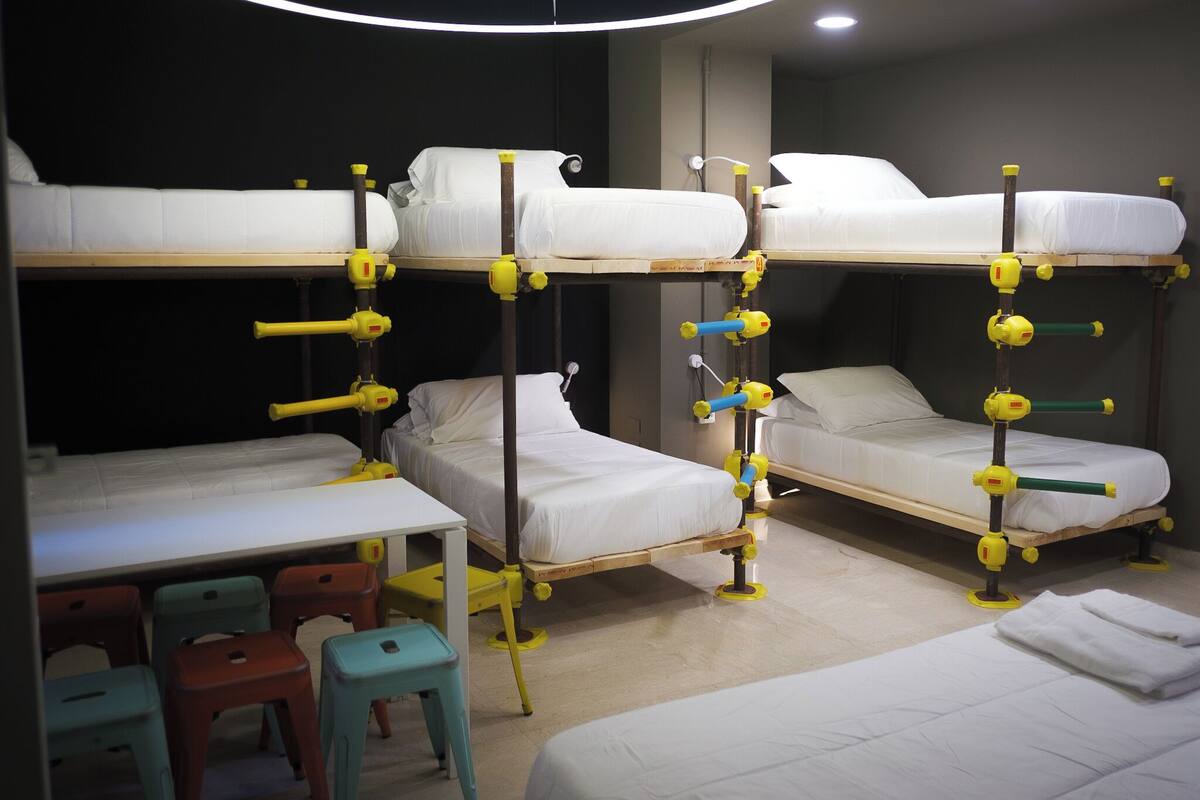 A dormitory room with bunk beds, a desk, and colorful stools.